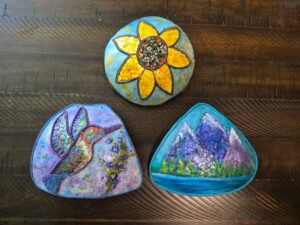 Three small decorated gourds sit on a table. One has a hummingbird in flight, one has a sunflower, and one has a scene with a lake in front of several mountains