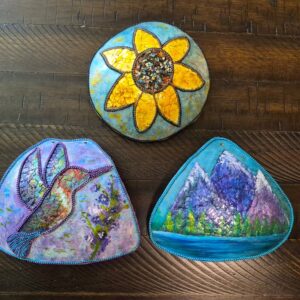 Three small decorated gourds sit on a table. One has a hummingbird in flight, one has a sunflower, and one has a scene with a lake in front of several mountains